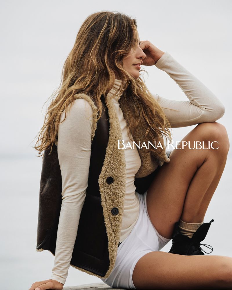 A woman models Banana Republic apparel outdoors, wearing a shearling-lined vest, long-sleeved shirt, shorts, socks, and boots against a cloudy sky.