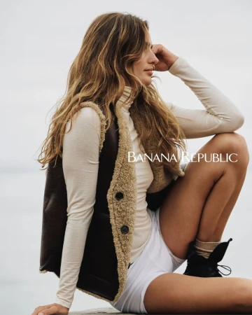 A woman models Banana Republic apparel outdoors, wearing a shearling-lined vest, long-sleeved shirt, shorts, socks, and boots against a cloudy sky.
