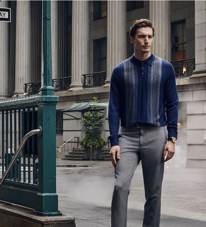 A stylish adult man stands in an urban setting wearing a blue striped sweater, grey pants, and a gold watch with an office building background.