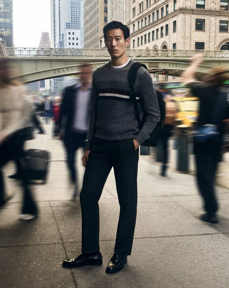 Asian man stands on city street in sweater and black pants with blurred pedestrians and buildings, creating a dynamic urban fashion image.