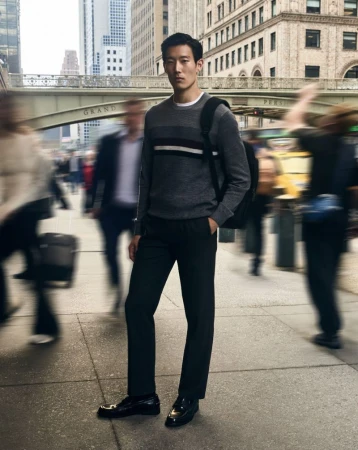 Asian man stands on city street in sweater and black pants with blurred pedestrians and buildings, creating a dynamic urban fashion image.