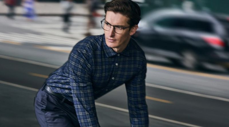Man in glasses wearing a blue plaid button-down shirt and dark trousers, leaning forward on a city street with blurry cars in the background.