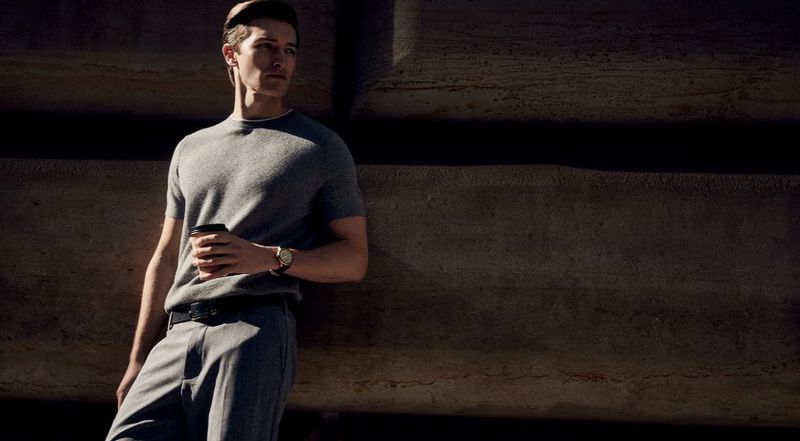 A man leans against a wall outside while holding a coffee cup, wearing a grey t-shirt and pants in a fashion style.