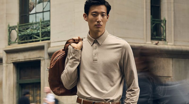 An Asian adult male poses outdoors with a brown leather backpack. He wears a long-sleeved polo shirt and a brown leather belt.