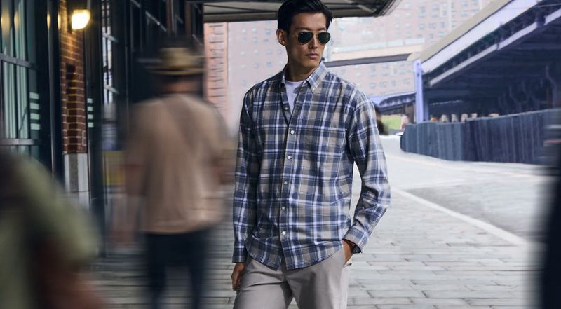 An Asian man in sunglasses and a blue and gray plaid shirt stands on a city street with blurred figures in the background.