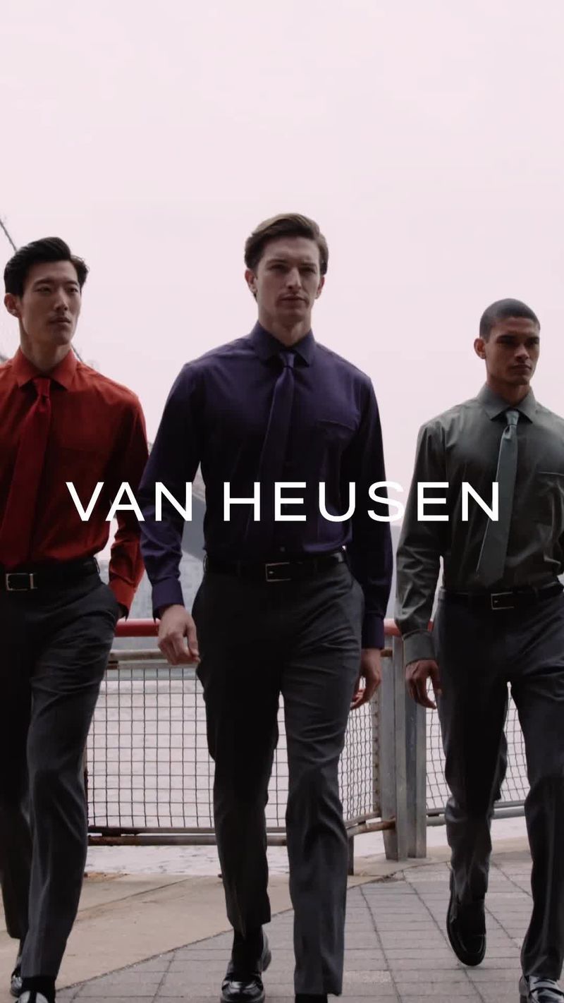 Three diverse adult men in fashionable formalwear walk outdoors against a muted urban backdrop promoting Van Heusen clothing.
