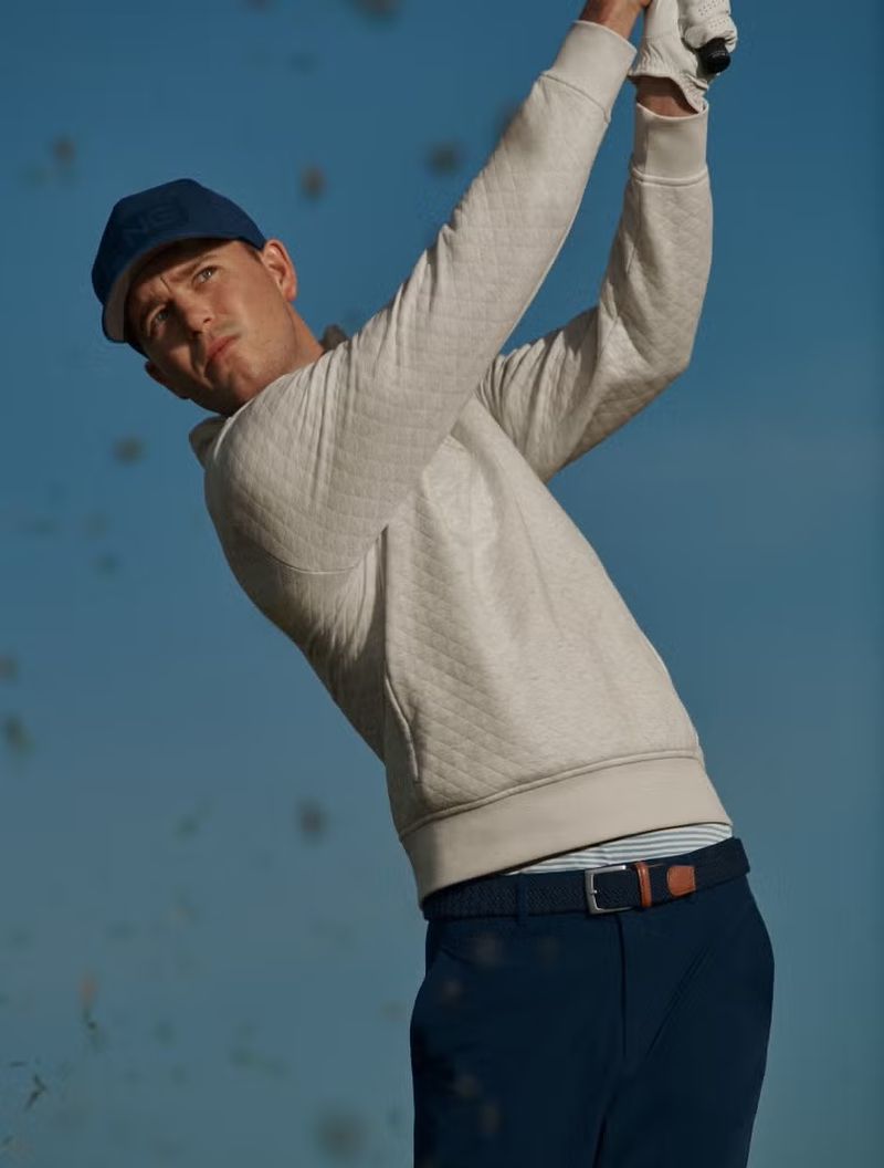 Man in a white sweater and navy hat swinging a golf club against a blue sky in a sports-themed outdoor shot, capturing athletic action.