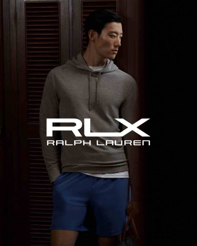 Asian male model in a grey hooded top and blue shorts poses in a dimly lit indoor setting. Ralph Lauren and RLX brand logos are prominent.