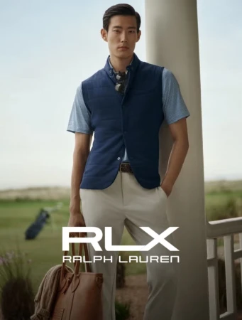 A stylish Asian man in a navy vest and tan pants poses on a golf course, holding a tote bag, in an advertisement for RLX Ralph Lauren.