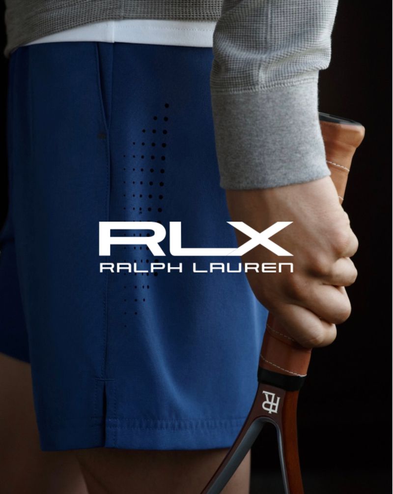 Athletic man holds a tennis racket. He wears blue athletic shorts and a gray sweatshirt.  RLX Ralph Lauren logo is featured.