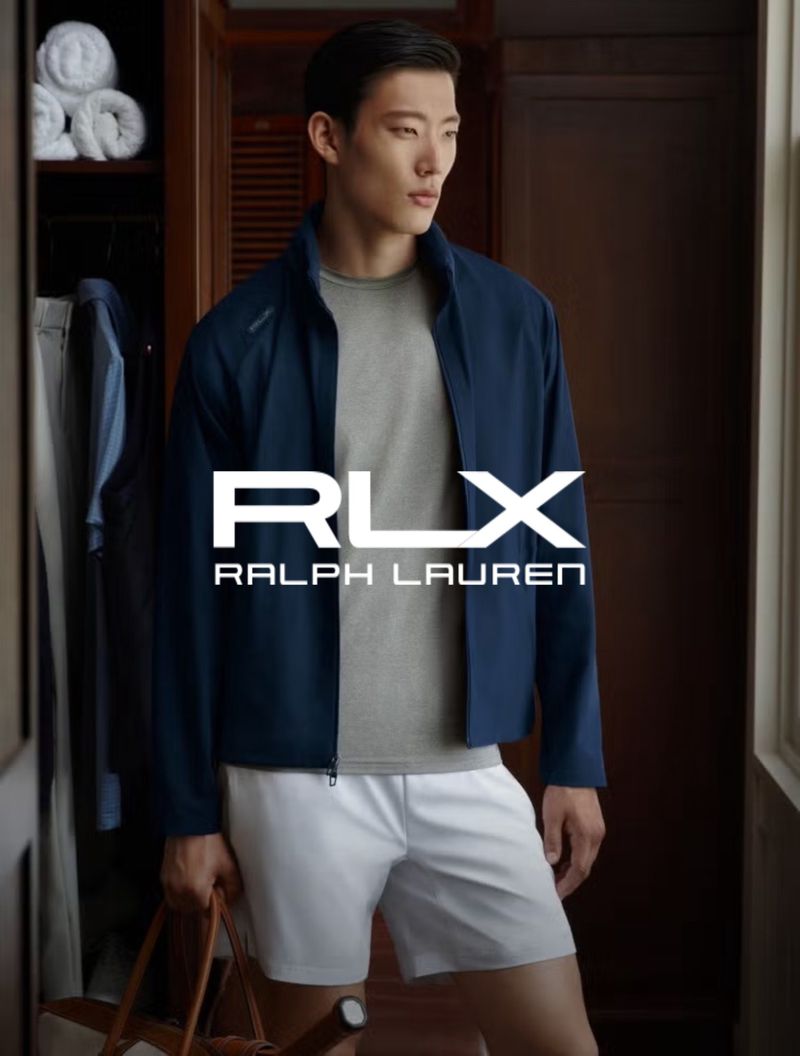 Asian male model in RLX Ralph Lauren advertisement, wearing a jacket and shorts, carrying a bag in an indoor setting.