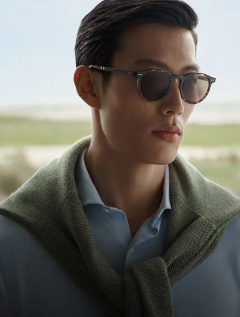 Close-up of an adult male model wearing sunglasses and a light blue shirt with a sage green sweater draped over his shoulders.