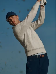 Model Juhyung Kang in a white golf sweatshirt and blue cap mid-swing against a clear blue sky background