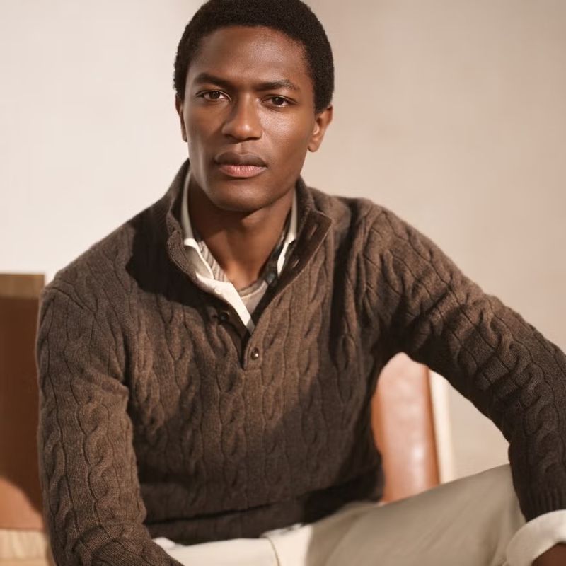Black man wearing a brown cable knit quarter zip sweater and light trousers seated indoors