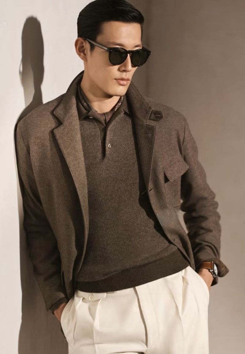 Portrait of a handsome Asian man wearing sunglasses, a brown polo shirt, a blazer, and white pants while leaning against a wall.