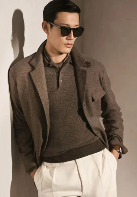Portrait of a handsome Asian man wearing sunglasses, a brown polo shirt, a blazer, and white pants while leaning against a wall.