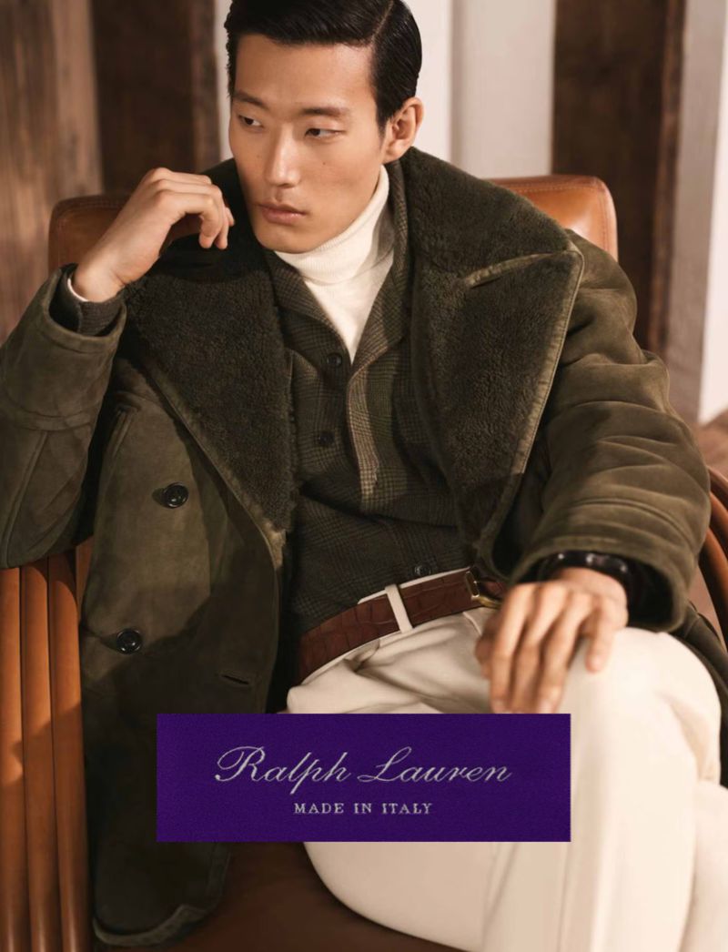 Asian male model sits in a chair wearing a dark green coat, tan turtleneck sweater, and off-white pants in a fashion advertisement.