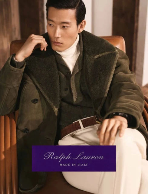Asian male model sits in a chair wearing a dark green coat, tan turtleneck sweater, and off-white pants in a fashion advertisement.
