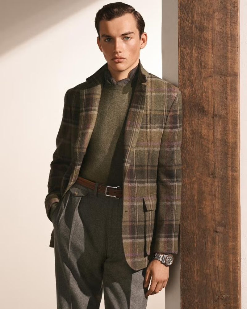 A young man in a plaid blazer, sweater, and grey pants stands near a wooden column in a studio, posing for a fashion shot.