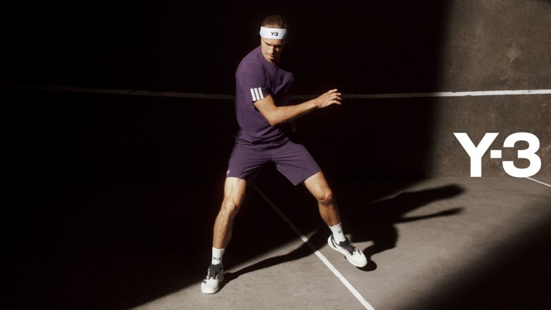 Male tennis player in purple Adidas Y-3 athletic wear on court in dynamic pose with a concrete background and dark, directional lighting.