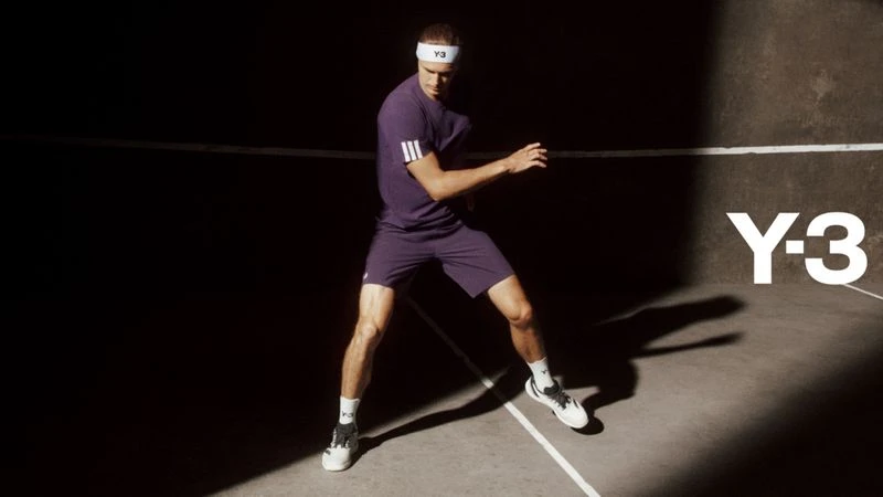 Male tennis player in purple Adidas Y-3 athletic wear on court in dynamic pose with a concrete background and dark, directional lighting.