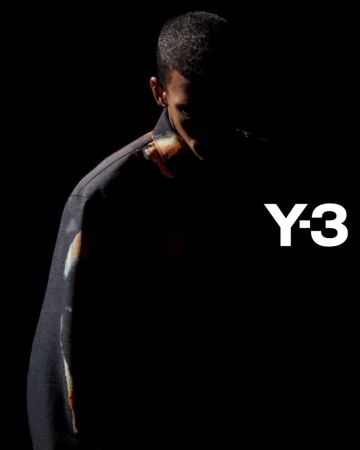 Dark, moody fashion shot featuring a man wearing a jacket, posed against a black background, highlighting the clothing and Y-3 logo.