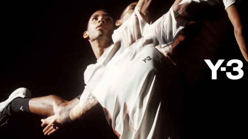 Two Black male adults in motion, illuminated against a dark background, wearing white t-shirts and black and white athletic shoes; Y-3 brand logo.