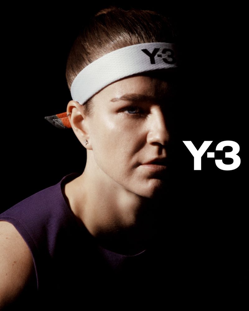 Close-up of a woman with a headband and purple tank top, facing forward in a dark, shadowed studio. The Y-3 logo is visible.