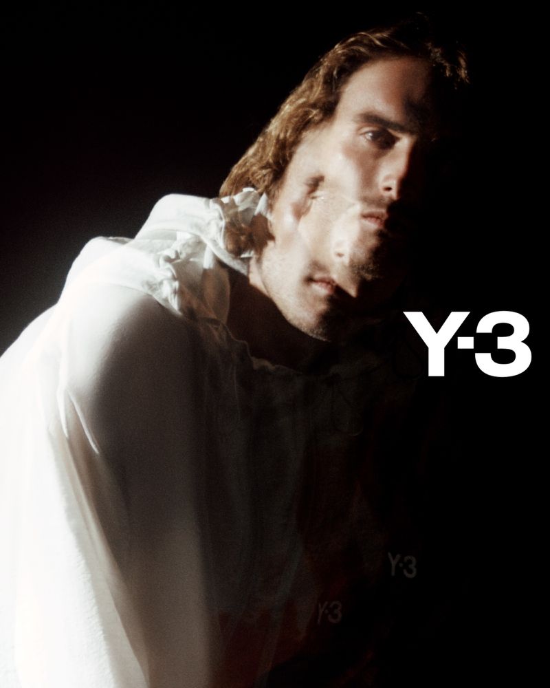 Fashion shot of a male model with long hair in a white shirt in front of a dark background with brand logo 'Y-3' visible.