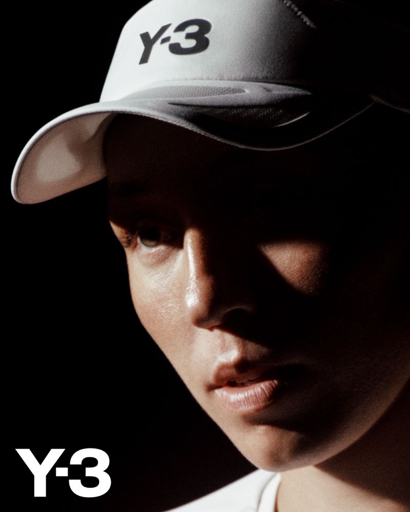Fashion shot of a model wearing a white baseball cap branded with the Y-3 logo, set against a black background, captured in a studio environment.