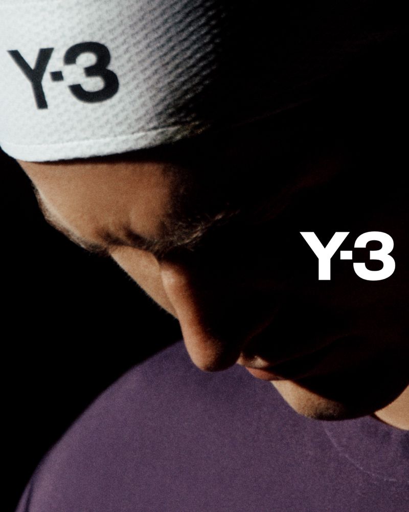 Close-up studio shot of an adult male wearing a white baseball cap with a black 'Y-3' logo and a purple t-shirt against a dark background.