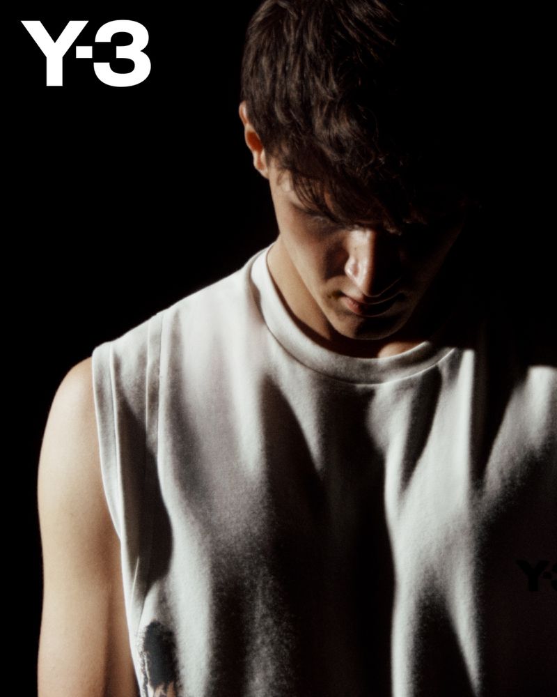Portrait of a young adult man in a white tank top against a dark background. The Y-3 logo is visible in the upper left corner.