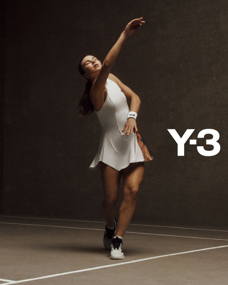Athletic woman in white tennis dress prepares to serve during a tennis match, indoors, highlighted by bright lighting, Y-3 brand visible.