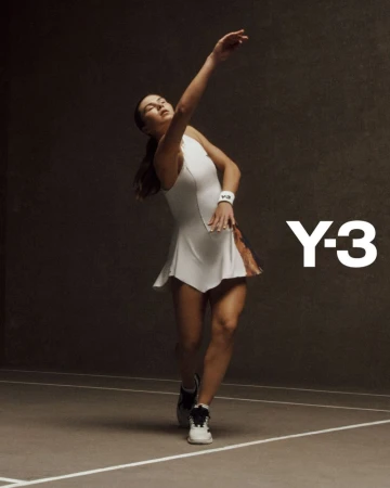Athletic woman in white tennis dress prepares to serve during a tennis match, indoors, highlighted by bright lighting, Y-3 brand visible.