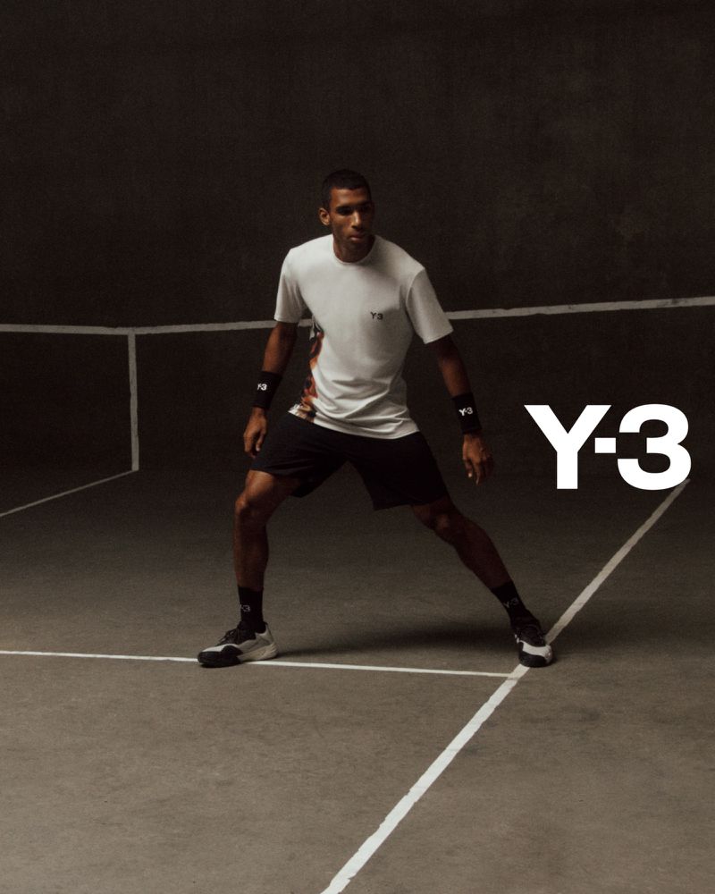 A man poses in athletic wear in a studio against a dark backdrop wearing a white shirt, black athletic shorts, and wristbands for Y-3 advertisement.