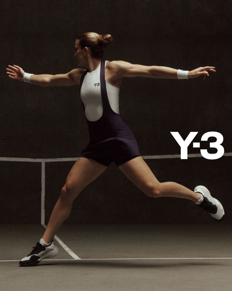 A woman wearing a white and navy athletic bodysuit and athletic shoes is in motion on an indoor court with a dark background.