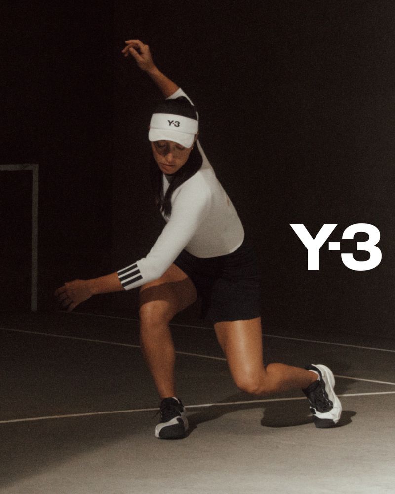 A woman wearing athleisure apparel swings a tennis racket. She is wearing a hat, long sleeve shirt, and a tennis skirt against a dark backdrop.