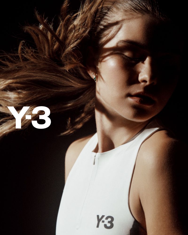 Close-up shot of a woman with brown hair in a white top, with the Y-3 logo visible, against a dark background with shadows.