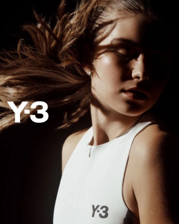 Close-up shot of a woman with brown hair in a white top, with the Y-3 logo visible, against a dark background with shadows.