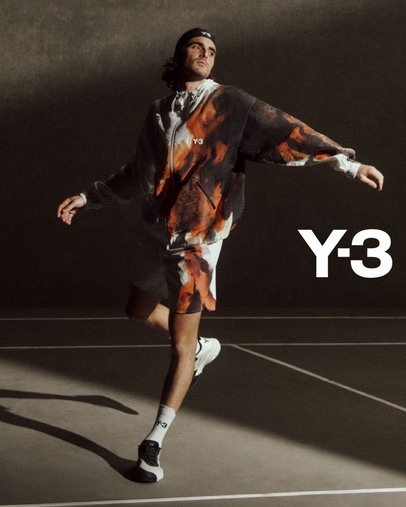 A man in a Y-3 printed hoodie and shorts poses dynamically on a neutral court with directional lighting, showcasing modern fashion.