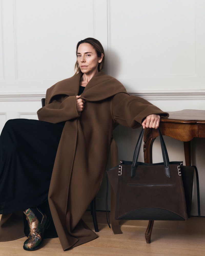 Woman seated in an oversized brown coat holding a large dark brown suede tote bag beside a wooden table against a white wall.