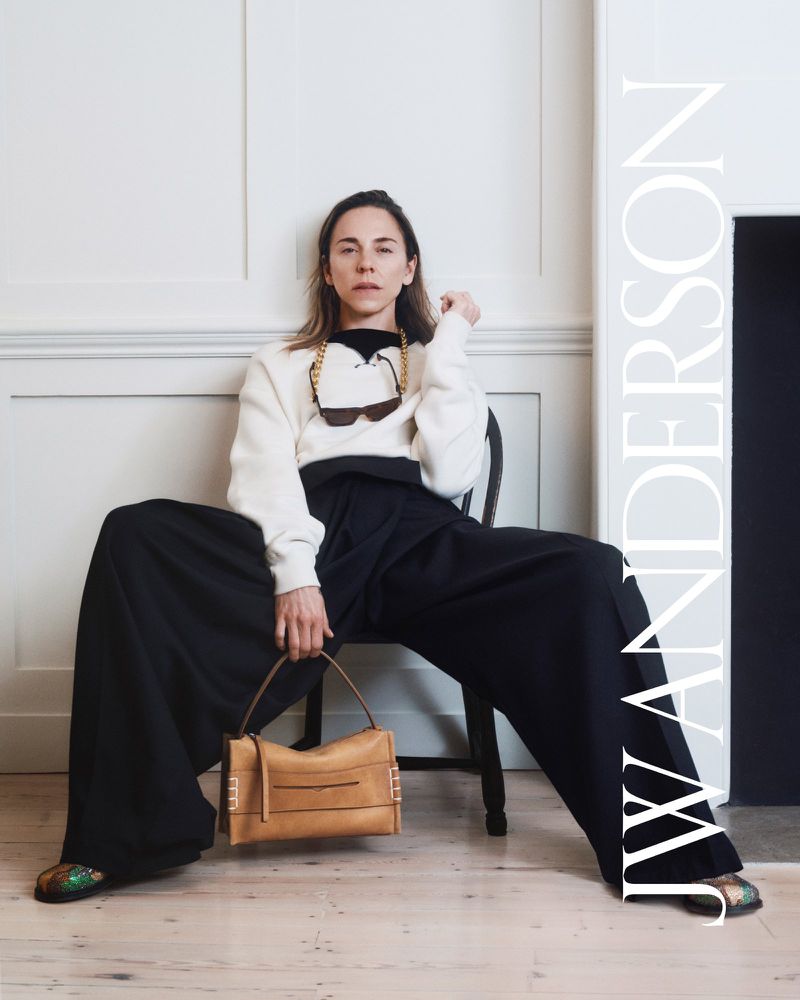 Woman seated on a chair in exaggerated wide-leg black trousers and a cream sweater, holding a tan leather J.W. Anderson Loafer bag.