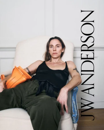 Adult female model poses in an off-white armchair with an orange handbag; she wears a black tank top and olive-green trousers indoors.