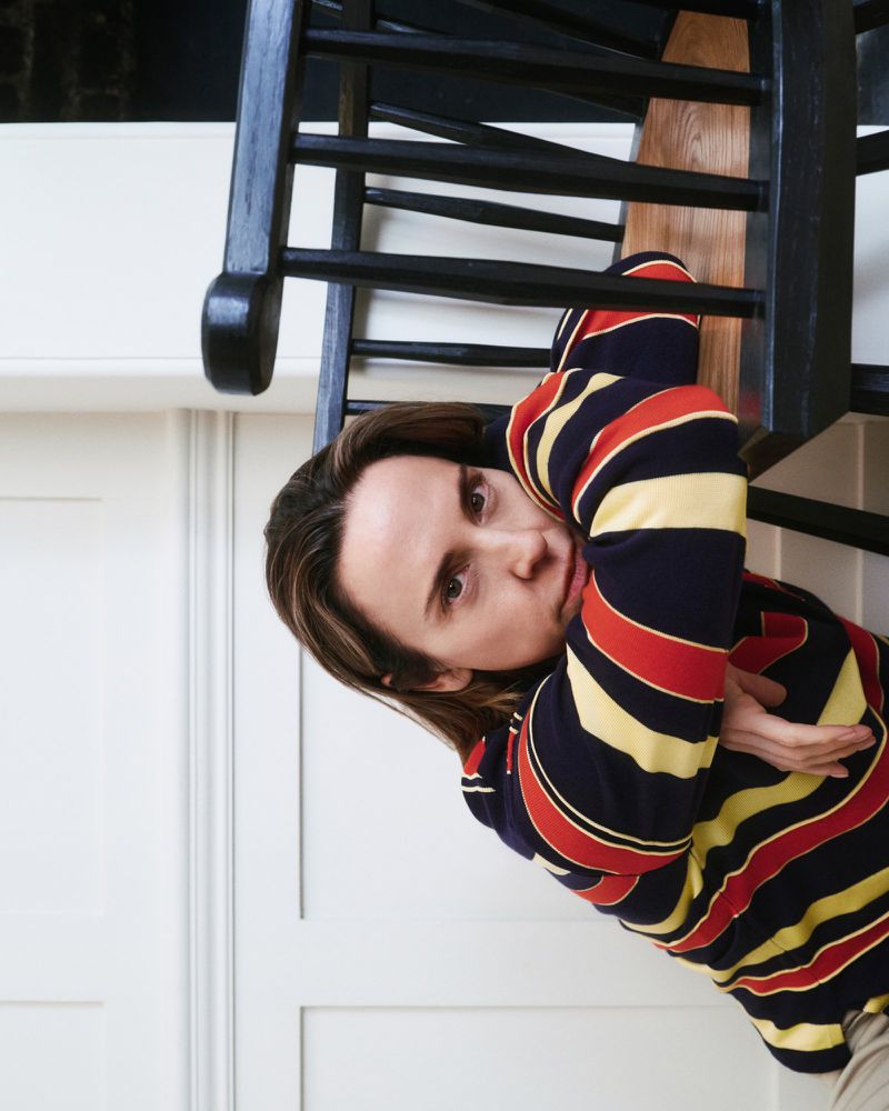 Portrait of a woman with medium-length brown hair, wearing a striped shirt, posing indoors near a black staircase and white walls.