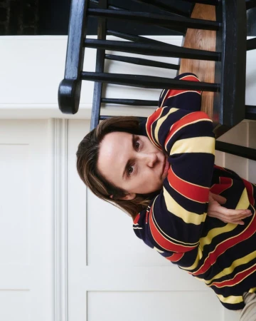 Portrait of a woman with medium-length brown hair, wearing a striped shirt, posing indoors near a black staircase and white walls.