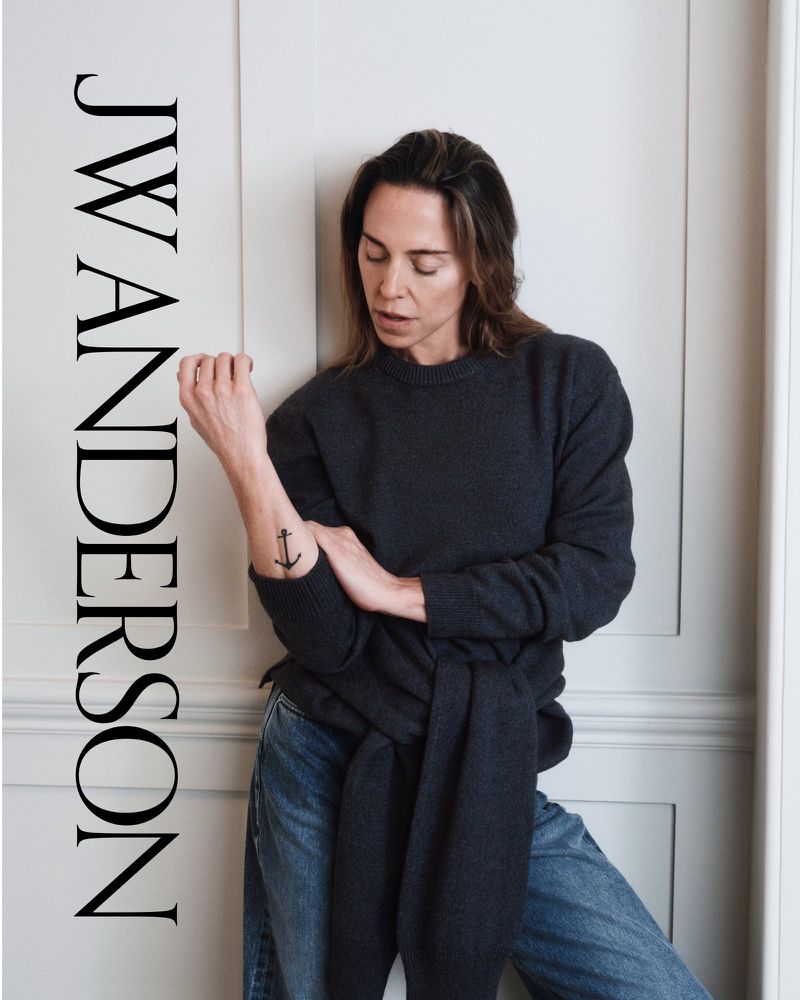 A woman in a grey sweater and blue jeans stands indoors against a white wall, with the text JW Anderson vertically on the left.