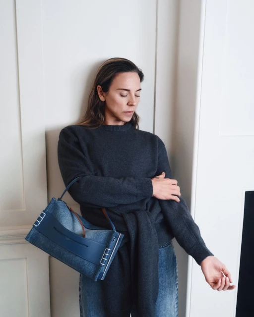 Model posing against white wall paneling with a blue denim JW Anderson Loafer Bag, wearing a dark charcoal knit sweater.
