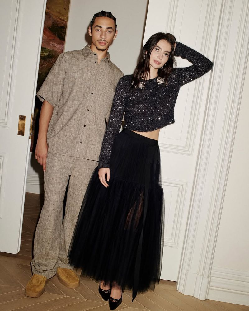 Two fashion models pose indoors wearing coordinated outfits: a tan linen suit and a sequined sweater with a black tulle skirt. 