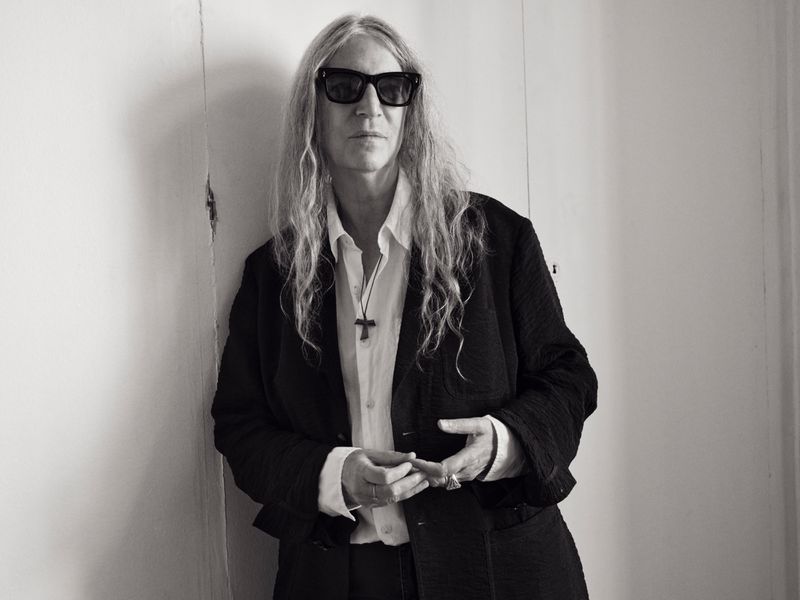 Black and white portrait of Patti Smith wearing sunglasses, blazer, and a cross necklace, standing against a white wall.