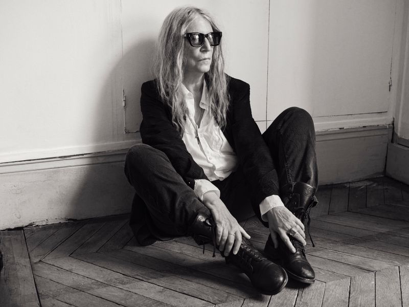 Patti Smith wearing dark eyewear, a black jacket, white shirt, and boots while seated on a chevron wood floor.